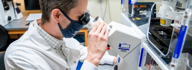 Researcher in white coat sitting in a lab looking through a microscope