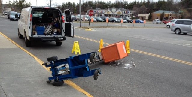 A blue and orange lift fell over into the street, yellow safety cones and a white van are stationed around it.