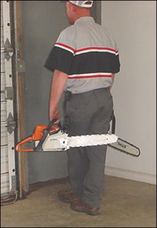 Person carrying chain saw at their side, chain bar is pointed behind them and the chain brake is engaged