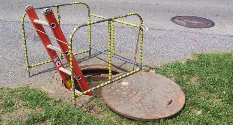 Pothole in the street is opened up with a ladder entering into it and yellow and black striped protective railing surrounds the area